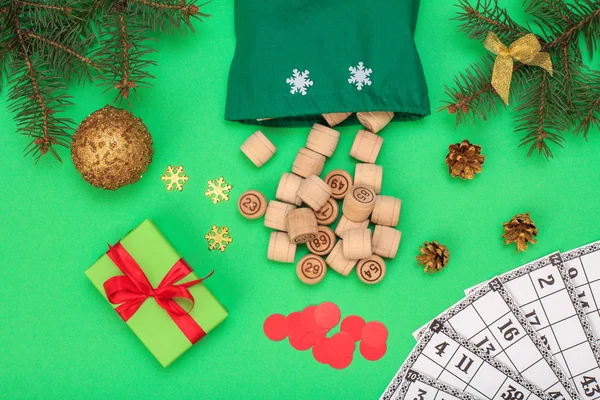 Board game lotto. Wooden lotto barrels with bag, game cards and red chips for a game in lotto, Christmas fir tree branch, cone, toy ball and gift boxes on green background. Top view