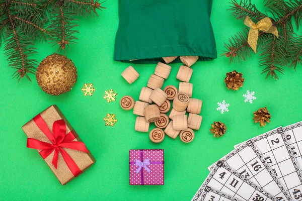 Board game lotto. Wooden kegs with bag and game cards for a game in lotto, Christmas fir tree branches, cones, toy ball, snowflackes and gift boxes on green background. Top view