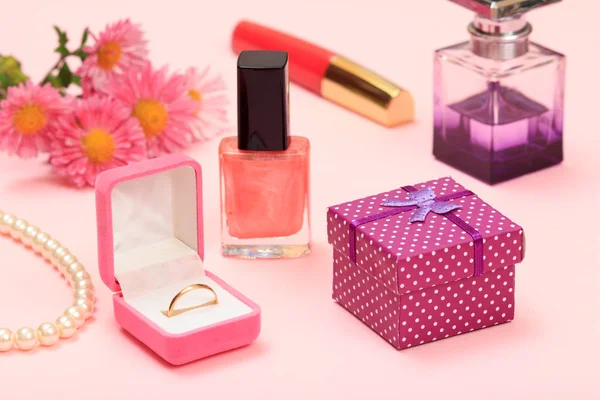 Gift box, flowers, bottles with nail polish and perfume, lipstick, golden ring in a box, beads on a pink background. Women cosmetics and accessories.