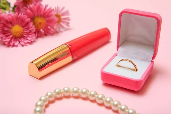 Golden ring in box, beads, lipstick and flowers on a pink background. Women jewelry and accessories.