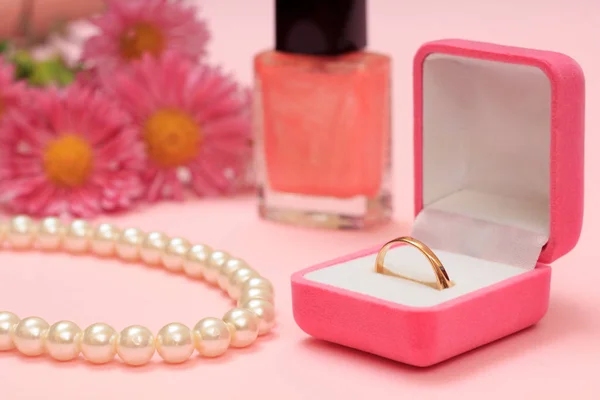 Golden ring in box, nail polish, beads and flowers on a pink background. Women jewelry and accessories.