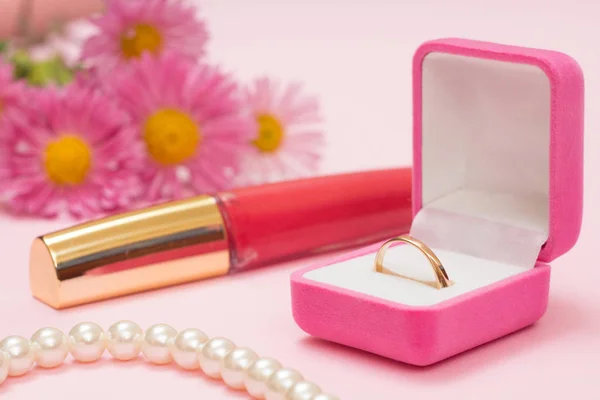 Golden ring in box, beads, lipstick and flowers on a pink background. Women jewelry and accessories.