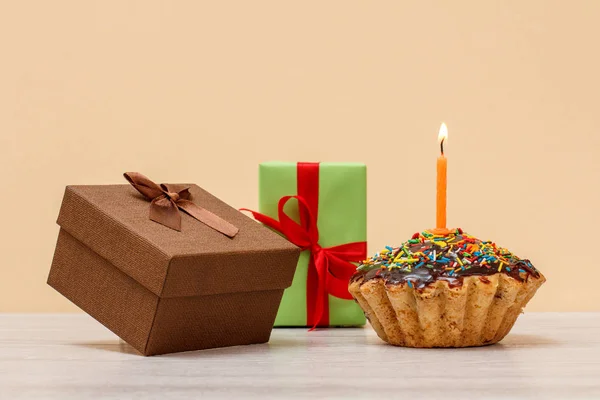 Tasty birthday muffin with chocolate glaze and caramel, decorated with burning festive candle and gift boxes on beige color background. Happy birthday minimal concept.