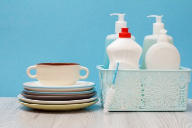 Plastik şişe bulaşık deterjanı, cam ve döşemedeki sepeti, temiz tabak temiz ve mavi arka plan üzerinde kase. Yıkama ve Temizleme kavramı.