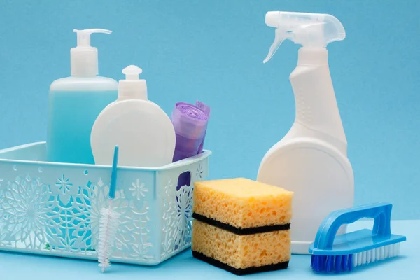 Plastic bottles of dishwashing liquid, glass and tile cleaner, garbage bags in basket, sponges and brush on blue background. Washing and cleaning concept.
