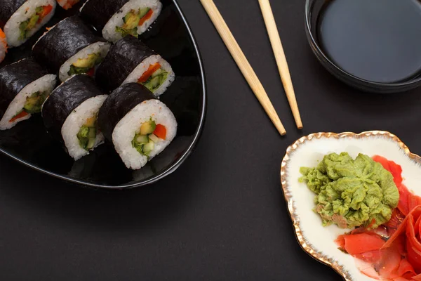 Sushi rolls in nori seaweed sheets with avocado and red fish on ceramic plate. Plate with red pickled ginger and wasabi. Bowl with soy sauce and wooden sticks. Top view.