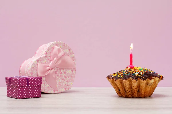 Gift boxes and tasty birthday muffin with chocolate glaze and caramel, decorated with burning festive candle on lilac background. Happy birthday minimal concept.