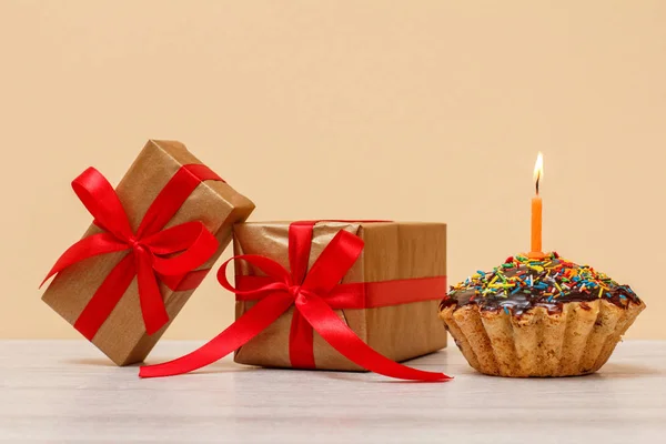 Tasty birthday muffin with chocolate glaze and caramel, decorated with burning festive candle and gift boxes on beige background. Happy birthday minimal concept.