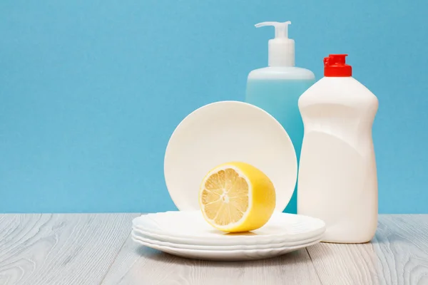 Plastic bottles of dishwashing liquid, glass and tile cleaner, clean plates and lemon on blue background. Washing and cleaning concept.