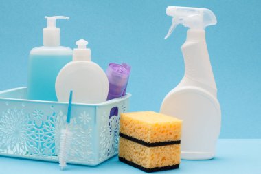 Plastic bottles of dishwashing liquid, glass and tile cleaner, garbage bags in basket, sponges and detergent for microwave ovens and stoves on blue background. Washing and cleaning concept.