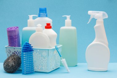Plastic bottles of dishwashing liquid, glass and tile cleaner, garbage bags in basket, brushes, sponge and detergent for microwave ovens and stoves on blue background. Washing and cleaning concept.