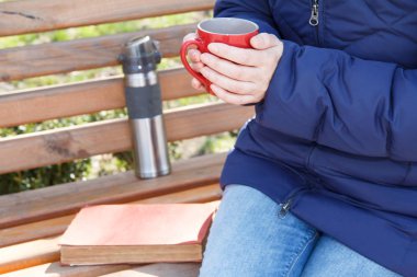 Kırmızı fincan kahve tutarak ve eski kitap ve termos bir bankta oturan kadın. Bayan park güneşli bir günde rahatlatıcı vardır. Bir tarih seçici odak.