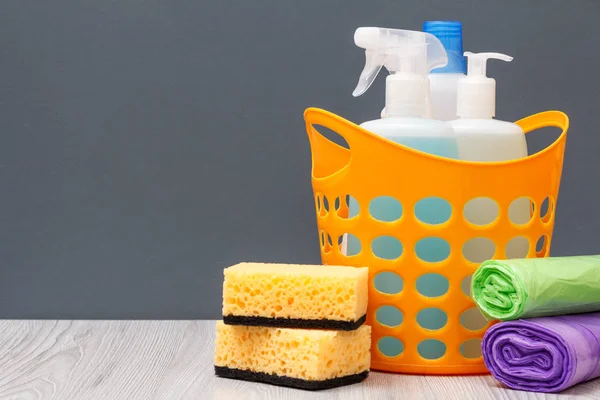 Plastic basket with bottles of dishwashing liquid and tile cleaner, detergent for microwave ovens and stoves, sponges and garbage bags on gray background. Washing and cleaning concept.