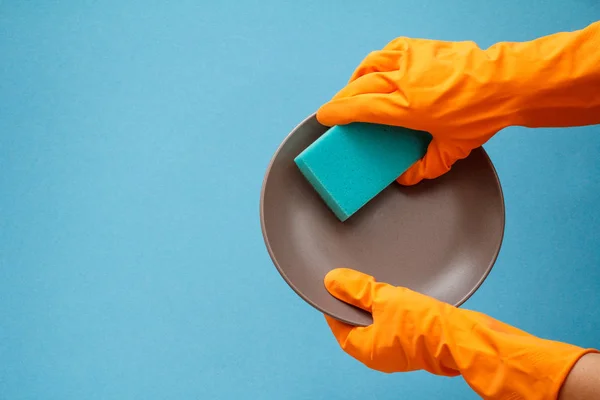 Women\'s hands in orange protective gloves with plate and sponge on blue background. Washing and cleaning concept.