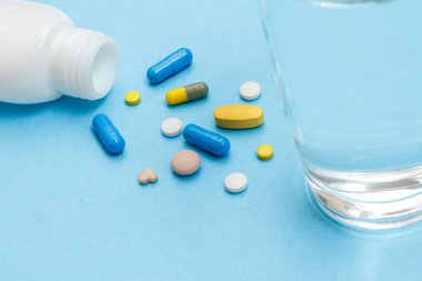 White bottle with different pills, capsules and glass of water on blue background.