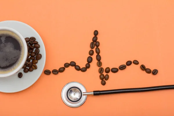 Cup of coffee on saucer with coffee beans folded in the form of a cardiogram and phonendoscope on peach background.