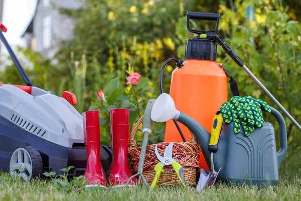 Rasaerba elettrico, stivali di gomma da giardino rossi, piccolo rastrello, potatore, cesto di vimini, spatola, spruzzatore di plastica e annaffiatoio . — Foto Stock