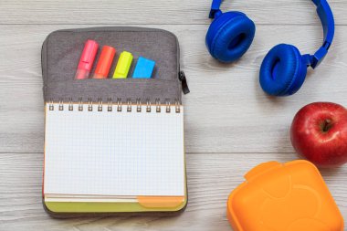 Gri arka plan üzerinde renkli keçekalem ve marker, sarı sandviç kutusu, elma ve kulaklık ile çanta-kalem durumda egzersiz kitabı.