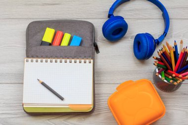 Gri arka plan üzerinde renkli keçekalem ve marker, sandviç kutusu, renkli kalemler ve kulaklıklar ile çanta-kalem durumda açık egzersiz kitabı.