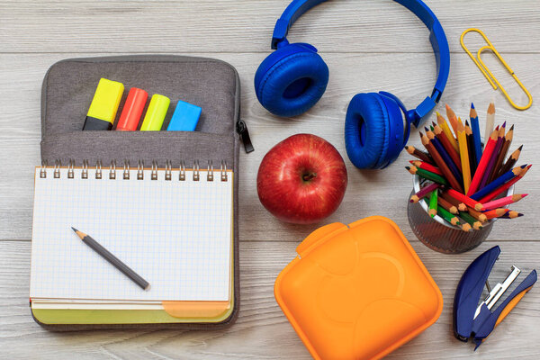 Lunch box, apple, headphones, open exercise book on bag-pencil case and school supplies on grey background.