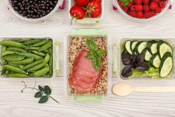 Plastic meal prep containers with boiled buckwheat porridge and slices of meat, fresh vegetables and berries.