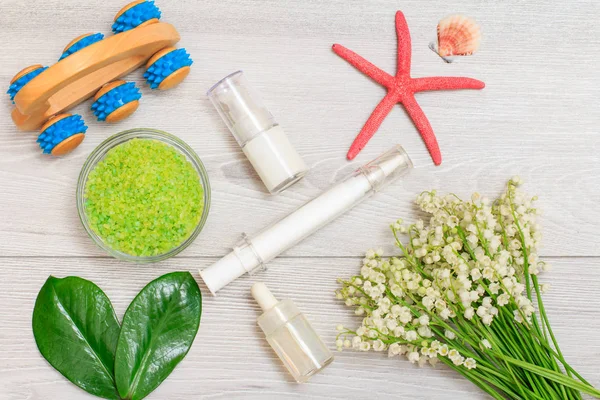 Syringe with eye cream, bottles with cream for face skin and aromatic oil, bowl with sea salt and bouquet of lilies of the valley.