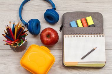 Okul malzemeleri... Elma, renkli kalemler, yemek kutusu, kulaklık ve gri ahşap panoları çanta-kalem durumda açık egzersiz kitabı.