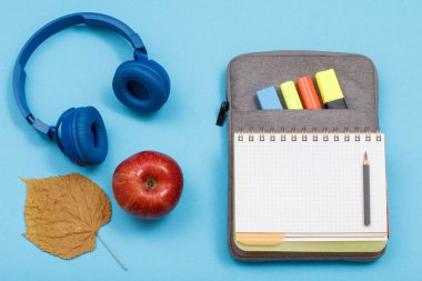 Elma, kulaklıklar, kuru yaprak, mavi arka plan üzerinde renk keçe kalem ve marker ile çanta-kalem durumda açık egzersiz kitabı.