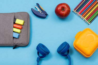 Öğle yemeği kutusu, renkli kalemler, elma, kulaklık, elma ve mavi arka plan üzerinde renk keçe kalem ve marker ile çanta-kalem durumda