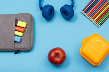 Öğle yemeği kutusu, renkli kalemler, elma, kulaklık, elma ve mavi arka plan üzerinde renk keçe kalem ve marker ile çanta-kalem durumda