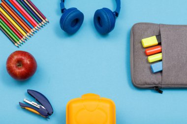 Öğle yemeği kutusu, renkli kalemler, elma, kulaklık, elma ve mavi arka plan üzerinde renk keçe kalem ve marker ile çanta-kalem durumda