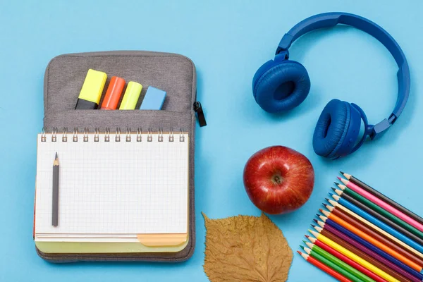 Color pencils, apple, headphones, open exercise book on bag-pencil case with color felt pens and marker on blue background.