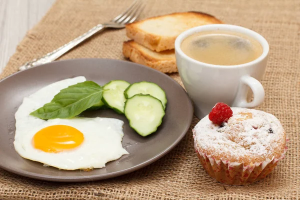 Bord met gebakken ei, vers gesneden komkommer en blad van basilicum, vork, kopje zwarte koffie, cupcake en toast. — Stockfoto
