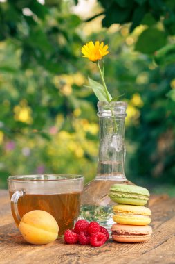 Olgun meyveler, cam şişede saplı calendula çiçeği, makaronlar ve bir fincan greeen çayı