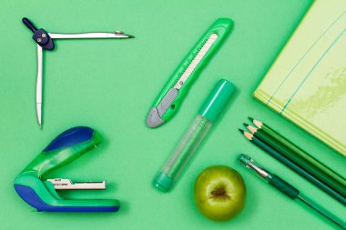 Yeşil arka plan üzerinde kitap, renkli kalemler, kalem, keçeli kalem, kağıt bıçak, pusula ve zımba.