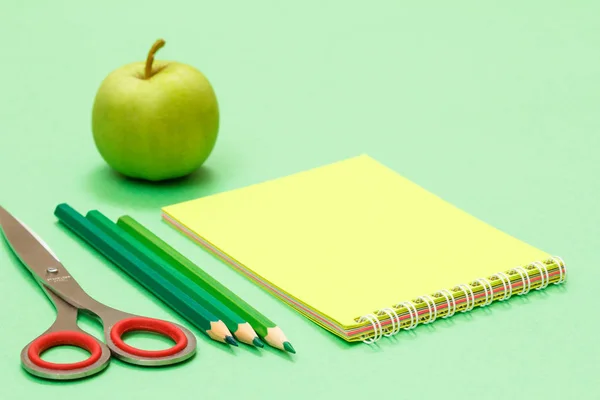 Nożyczki, kolorowe ołówki, Notatnik i jabłko na zielonym tle. — Zdjęcie stockowe