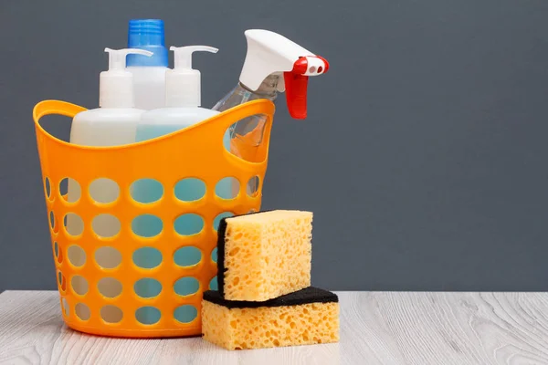 Bottles of dishwashing liquid and sponges in a basket on wooden desk.