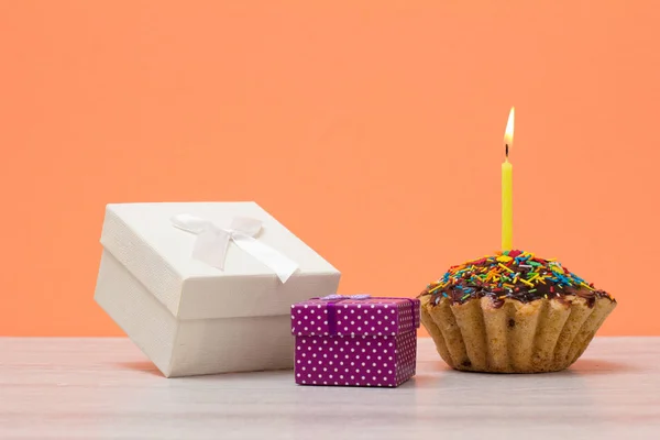 Sabroso panecillo de cumpleaños con vela festiva ardiente y cajas de regalo sobre fondo de melocotón . — Foto de Stock