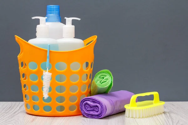 Bottles of dishwashing liquid, brush in a basket and garbage bags on gray background.