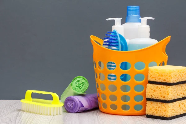 Bottles of dishwashing liquid, brush in a basket and sponges, brush, garbage bags on gray background.