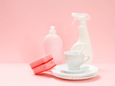 Bulaşık deterjanı şişeleri, süngerler, beyaz tabaklar, bir fincan ve pembe arka planda bir fincan. Yıkama ve temizlik konsepti.