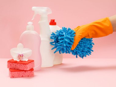Plastik çamaşır şişeleri, süngerler ve pembe arka planda paspas tutan kauçuk eldivenli bir el. Yıkama ve temizleme seti.