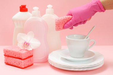 Plastik koruyucu eldivenli bir kadın eli, sünger, beyaz tabaklar, bir fincan, pembe arka planda bir fincan. Yıkama ve temizlik konsepti.