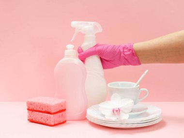Plastik koruyucu eldivenli bir kadın, bir şişe çamaşır suyu, beyaz tabaklar, bir fincan, pembe arka planda bir fincan. Yıkama ve temizlik konsepti.