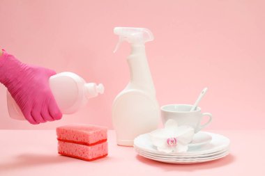 Kadın eli nitril koruyucu bir eldiven içinde bir şişe bulaşık deterjanı, beyaz tabaklar, bir fincan, pembe arka planda bir fincan. Yıkama ve temizlik konsepti.