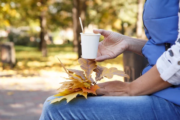 Young woman sitting in the city park and holding a cup of coffee and dry autumn leaves in her hands. Autumn composition.