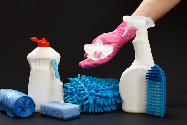 Female hands in nitrile gloves holding an orchid flower near the plastic bottles of washing liquid with a cleaning set on the black background. Washing and cleaning set.