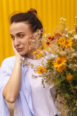 Güzel esmer kadınla sarı arka plan üzerinde topuz kafasındaki alanları çiçekler elinde tutarken mavi gömlek ve beyaz tişört giyiyor. Erkek çiçek getirmek