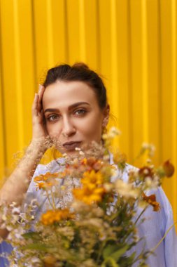 Güzel esmer kadınla sarı arka plan üzerinde topuz kafasındaki alanları çiçekler elinde tutarken mavi gömlek ve beyaz tişört giyiyor. Erkek çiçek getirmek