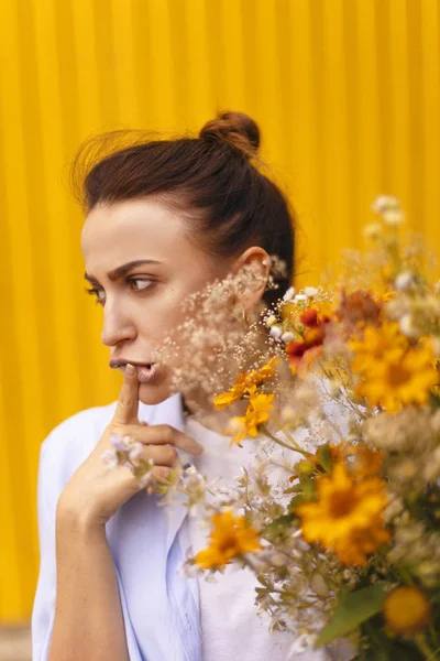 Beautiful brunette woman on yellow background with bun on her head wearing blue shirt and white t-shirt while holding fields flowers on her hand. Female bringing to flower — Stock Photo, Image